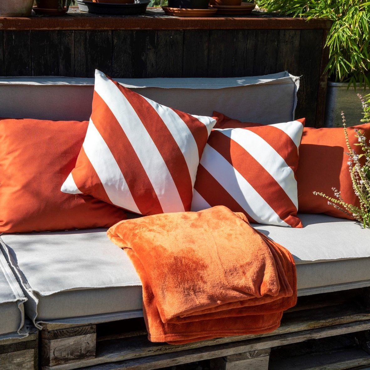 Terracotta sierkussen op tuinbank, met gestreept kussen ernaast en plaid