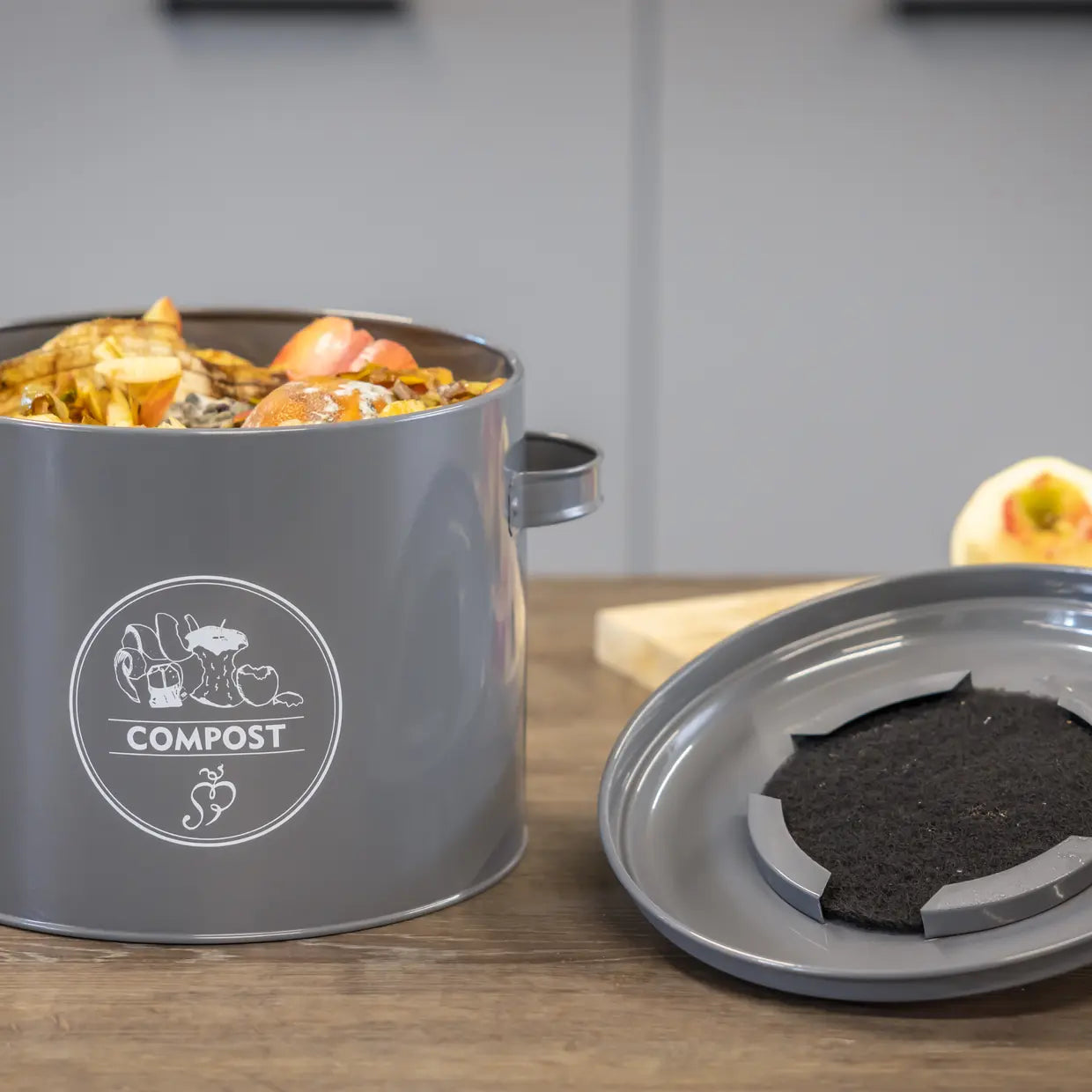 Keukencomposter antraciet met etensresten en losse bodemplaat met donkere compost.