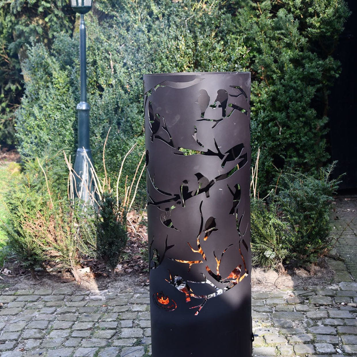 Cilindervormige vuurkorf met vogeluitsnedes op takken en vlammen