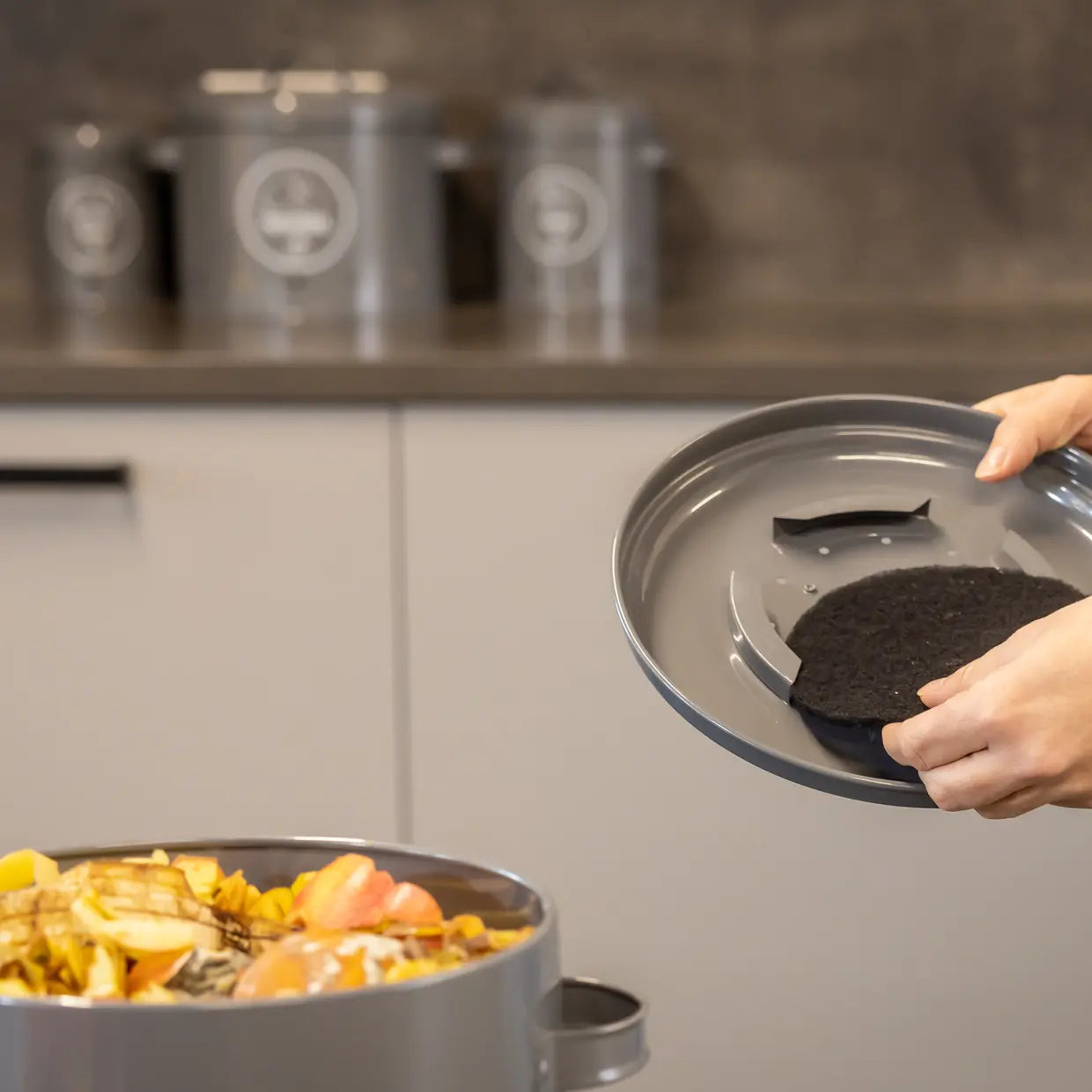 Keukencomposter antraciet met open bovenkant; hand legt donkere compost in de bak.